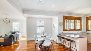 A spacious living room with a wooden floor, a dining area with a white table and chairs, and a kitchen with wooden cabinets and a bar area.