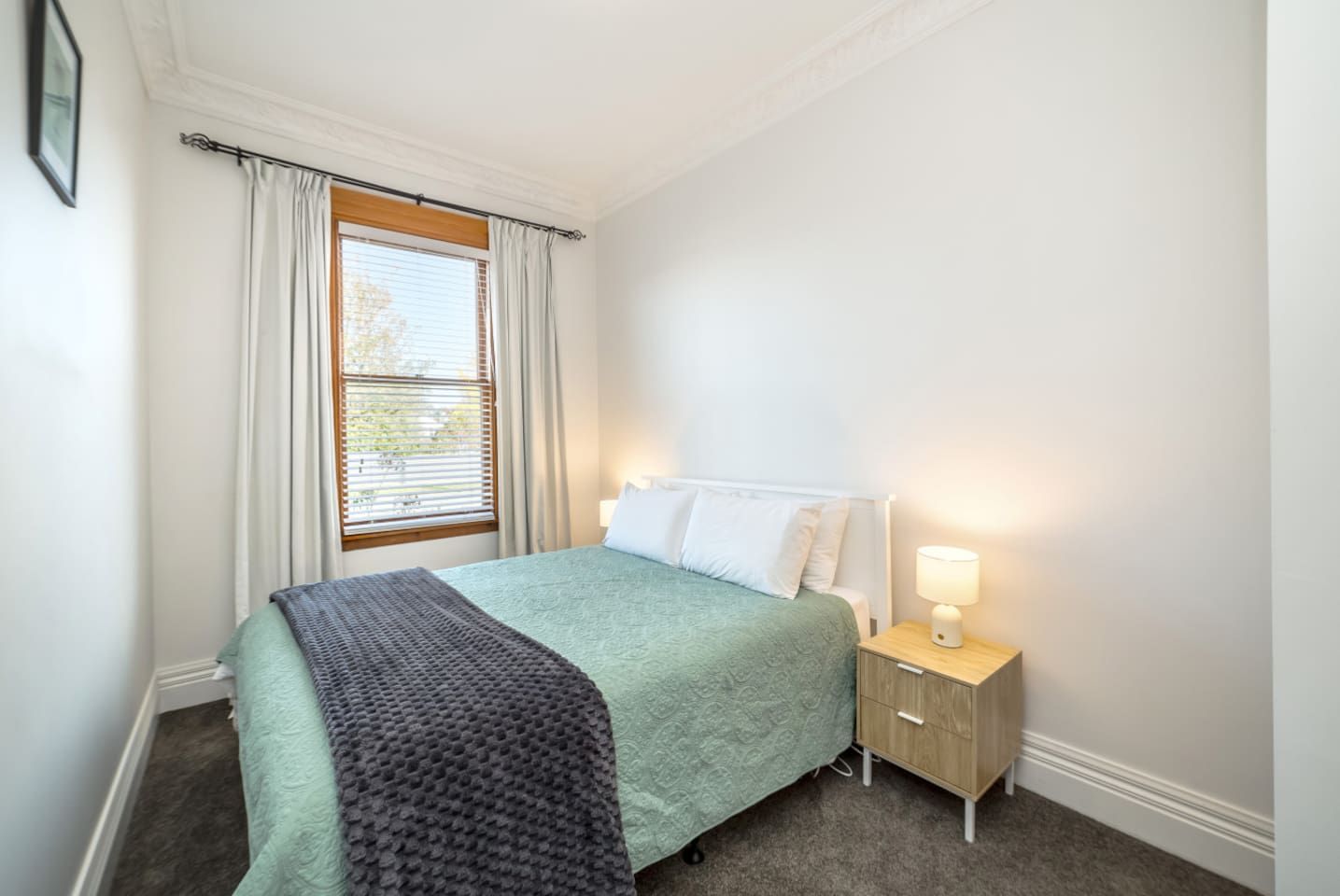 A bedroom with a bed, a bedside table with a lamp, a window with curtains, and a carpeted floor.