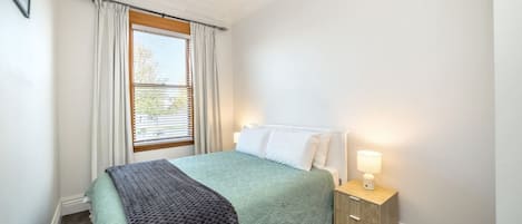A bedroom with a bed, a bedside table with a lamp, a window with curtains, and a carpeted floor.