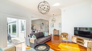 A spacious living room with a grey sofa, a round coffee table, a wicker chair, a TV stand, and a chandelier.