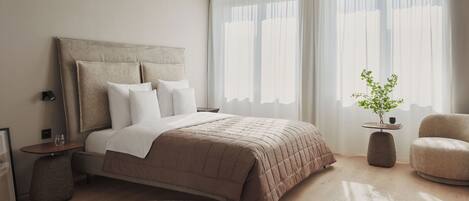 Een moderne slaapkamer met een groot bed, nachtkastjes, een klein bijzettafeltje, een stoel en een potplant.