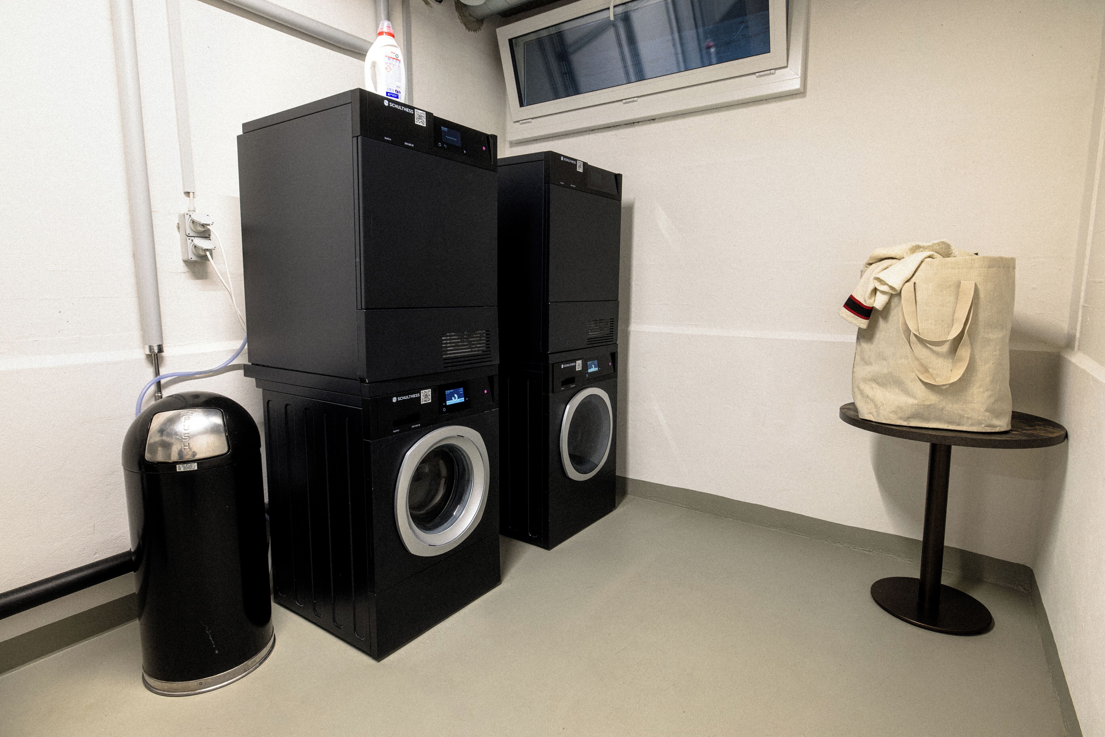 A laundry area with two black washing machines, a trash bin, and a small table with a bag on it.