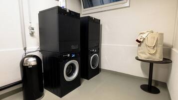 A laundry area with two black washing machines, a trash bin, and a small table with a bag on it.