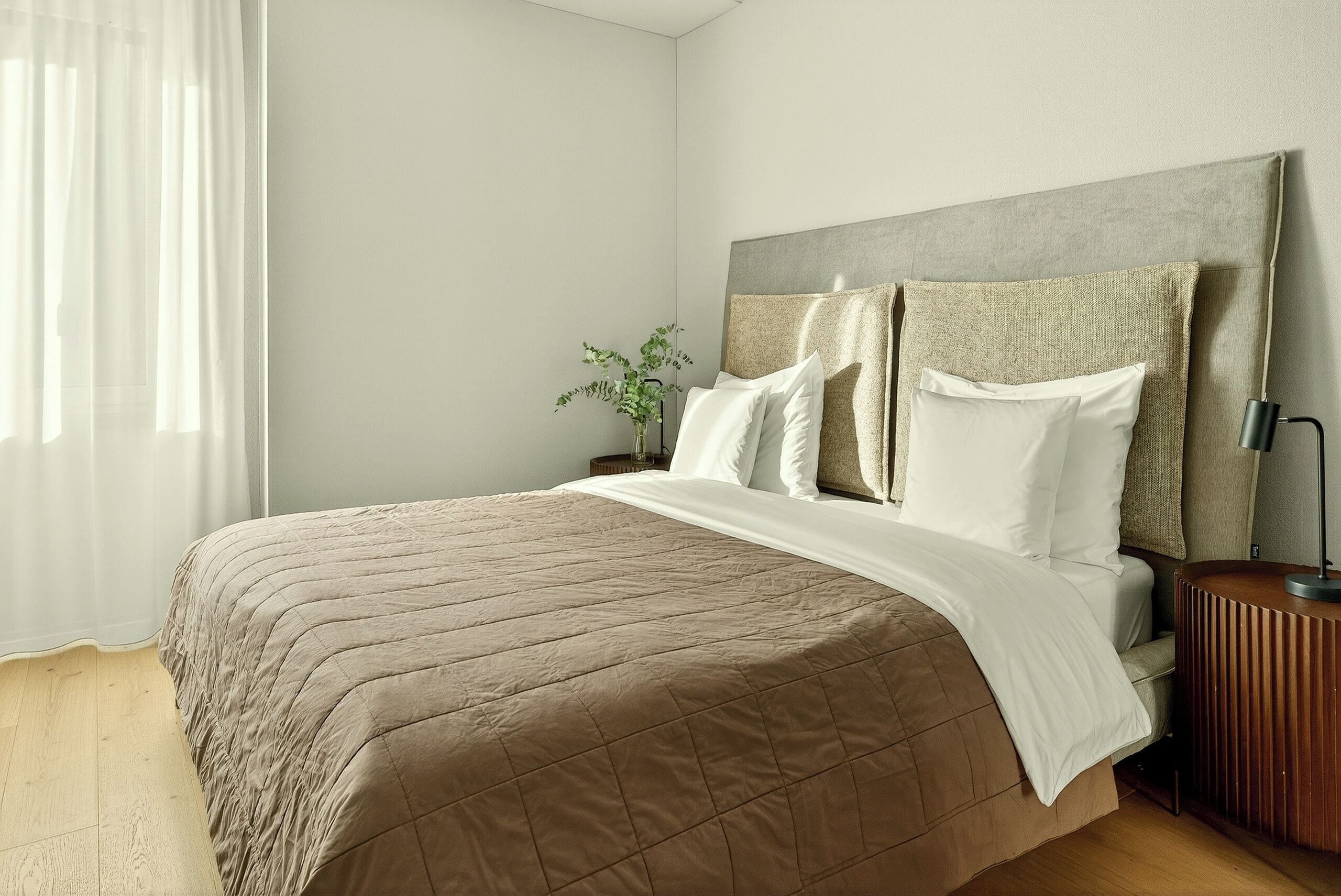 Een netjes opgemaakt bed met een beige deken, een grijs hoofdbord en een nachtkastje met een lamp.