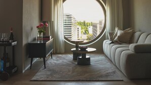 A modern living room with a round mirror, a sofa, a small table with a vase of flowers, and a view of buildings through a large window. - Belmont Residences (Opfikon)