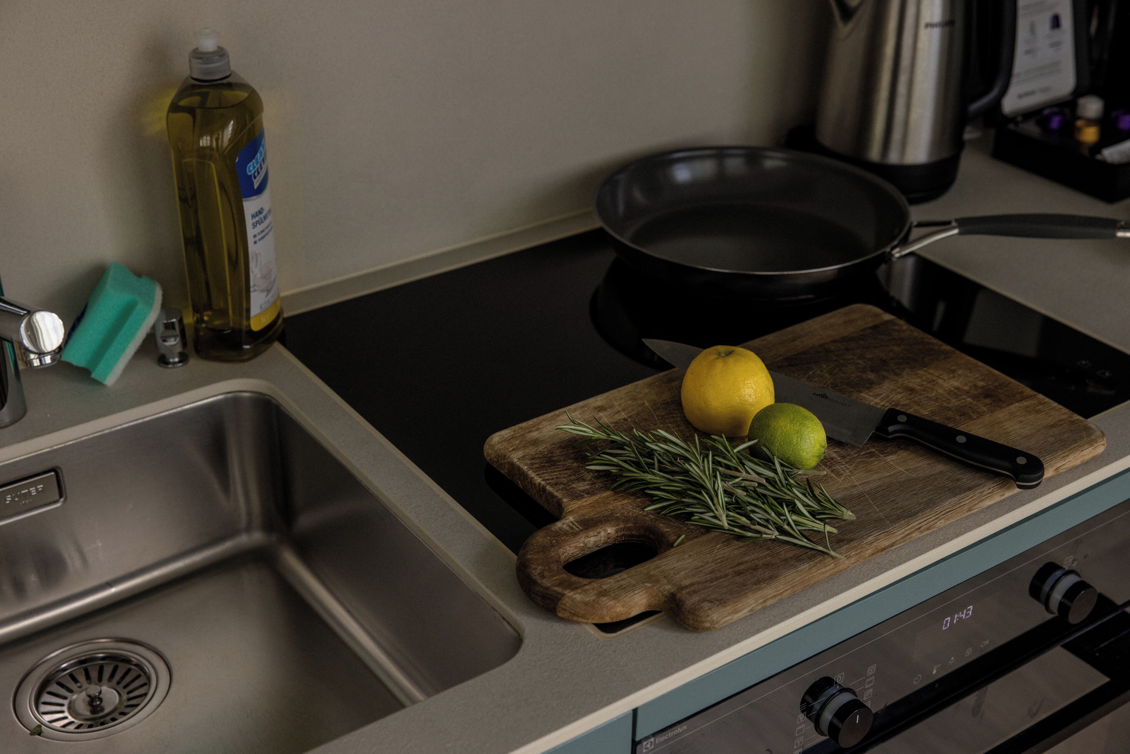 A kitchen with a sink, a cutting board with a lemon and rosemary, a frying pan, and a bottle of dish soap.