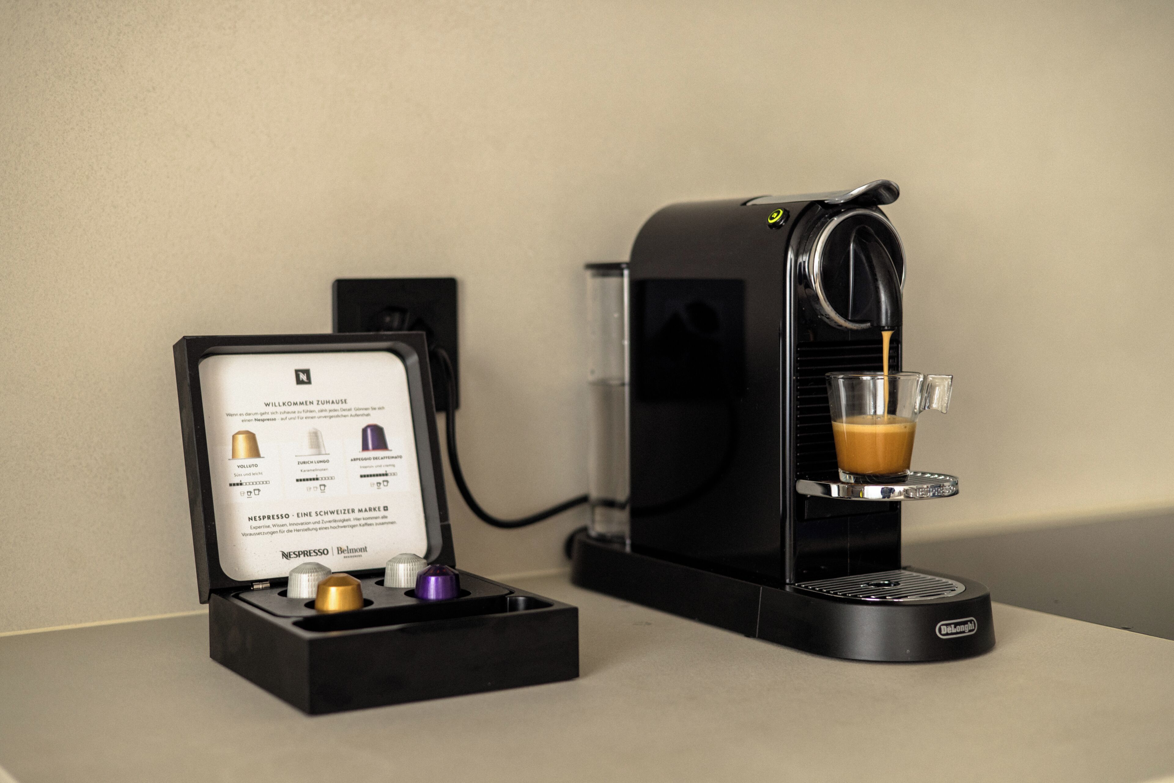 Eine Nespresso-Kaffeemaschine mit einer Glaskanne Kaffee, einem Nespresso-Kapselbehälter und einer wandmontierten Steckdose.