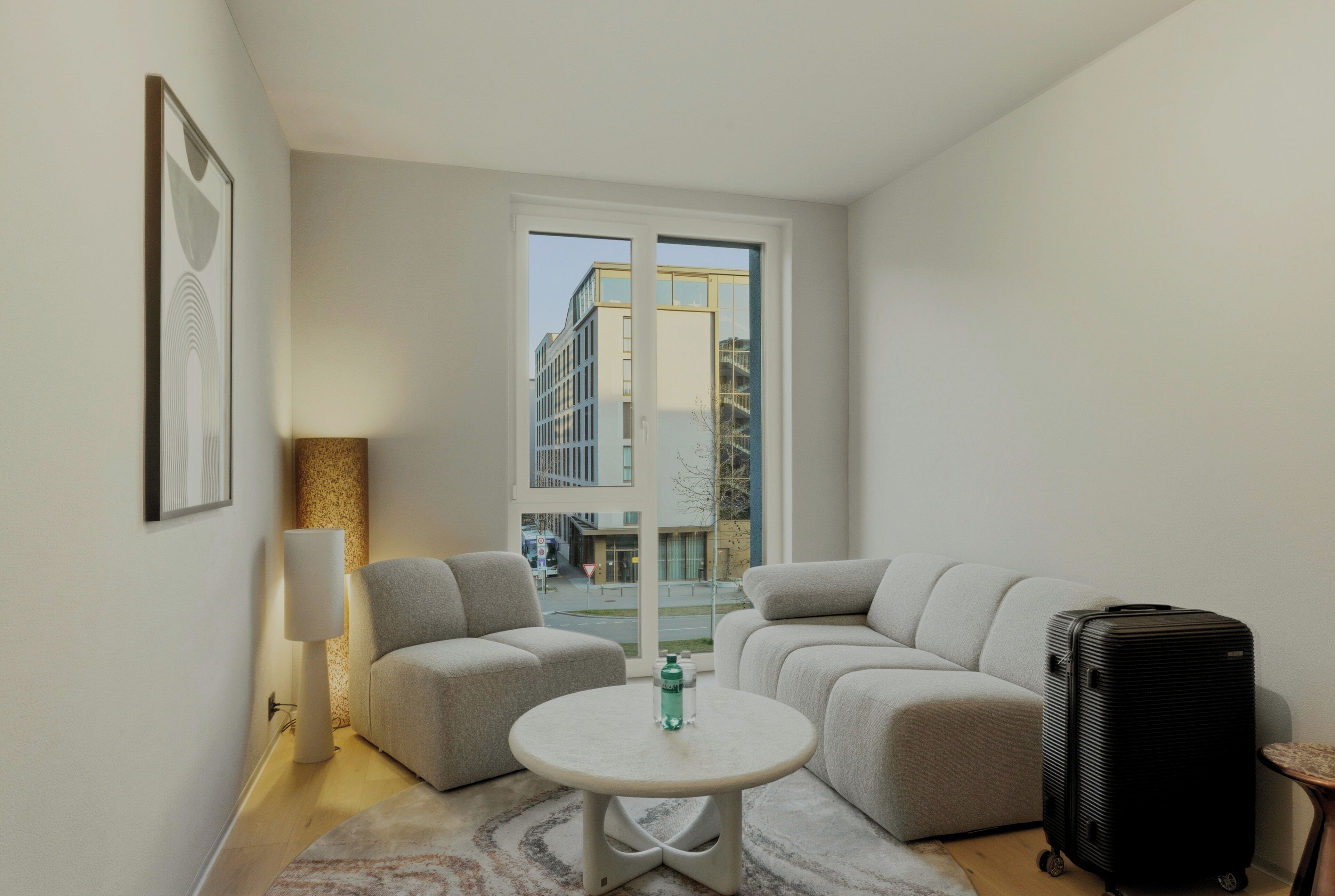 A modern hotel room with a sofa set, a small round table, a lamp, a suitcase, and a large window offering a view of the city.