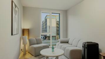 A modern hotel room with a sofa set, a small round table, a lamp, a suitcase, and a large window offering a view of the city.