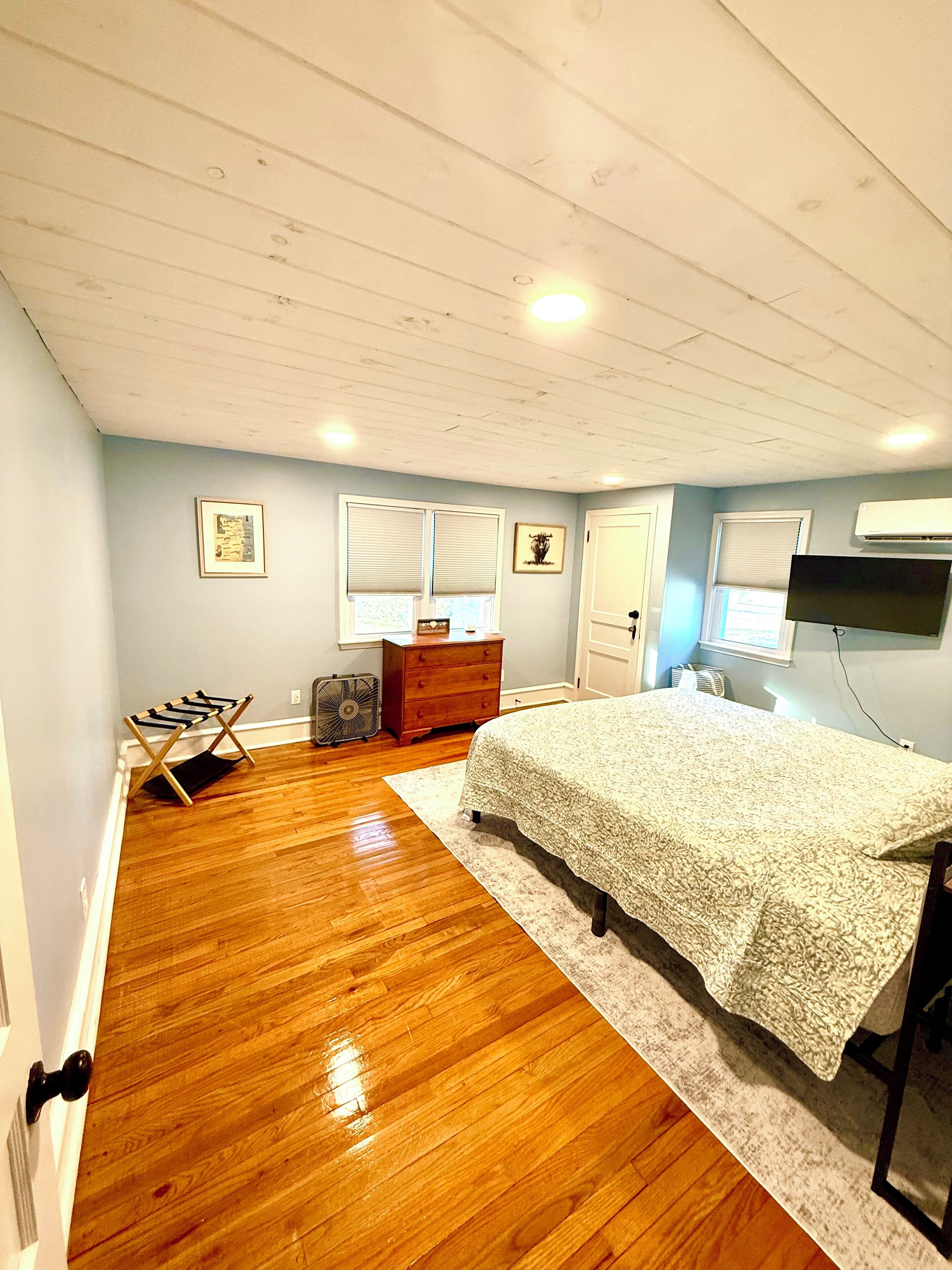 A bedroom with a wooden floor, a bed with a white bedspread, a dresser, a fan, and a wall-mounted TV.