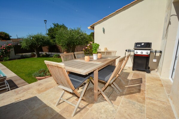 An outdoor patio with a wooden table and chairs, a barbecue grill, and a well-maintained lawn.