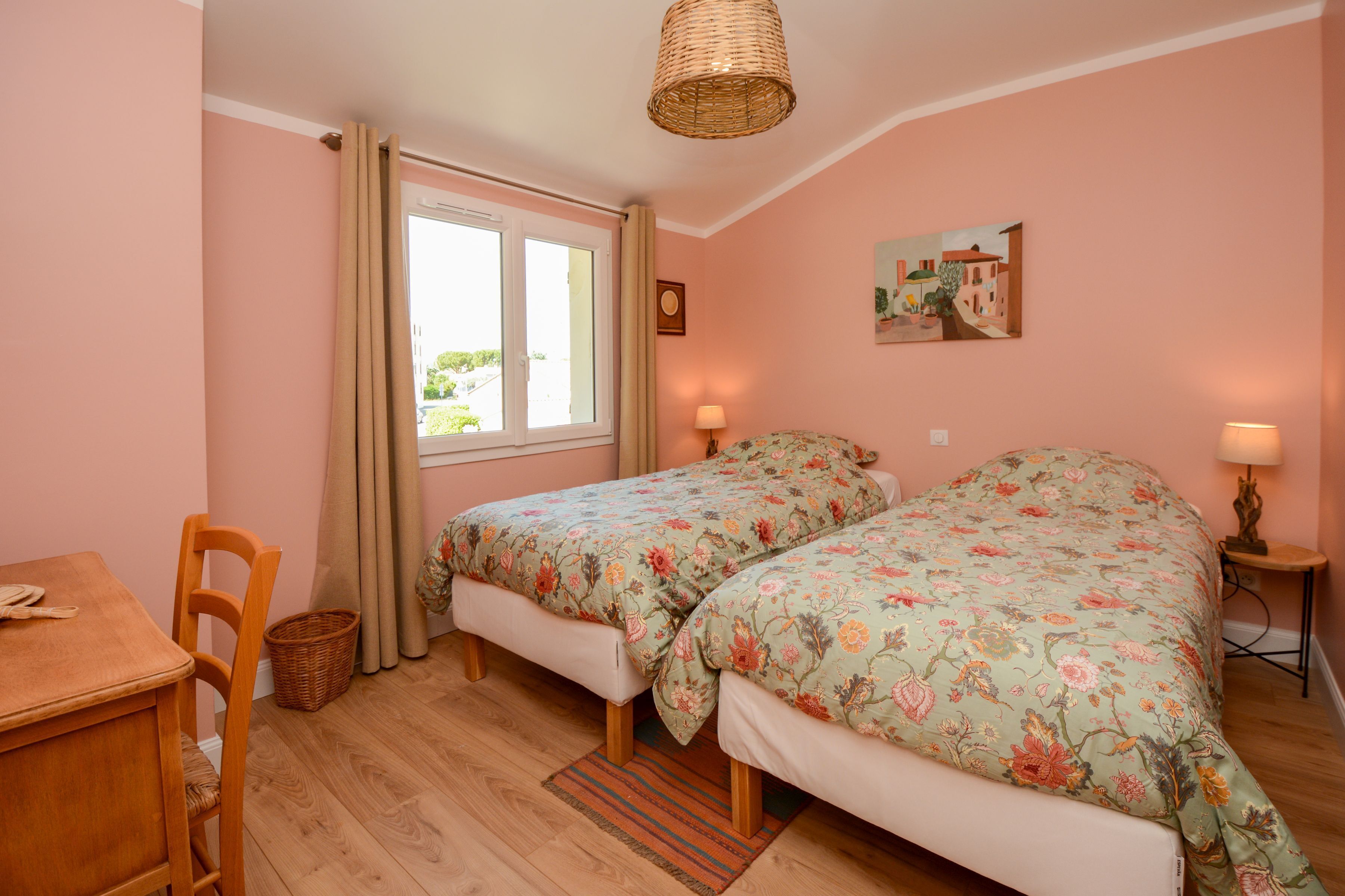 A bedroom with two beds, a wooden table, a chair, a window with curtains, a lamp on each side of the beds, and a painting on the wall.