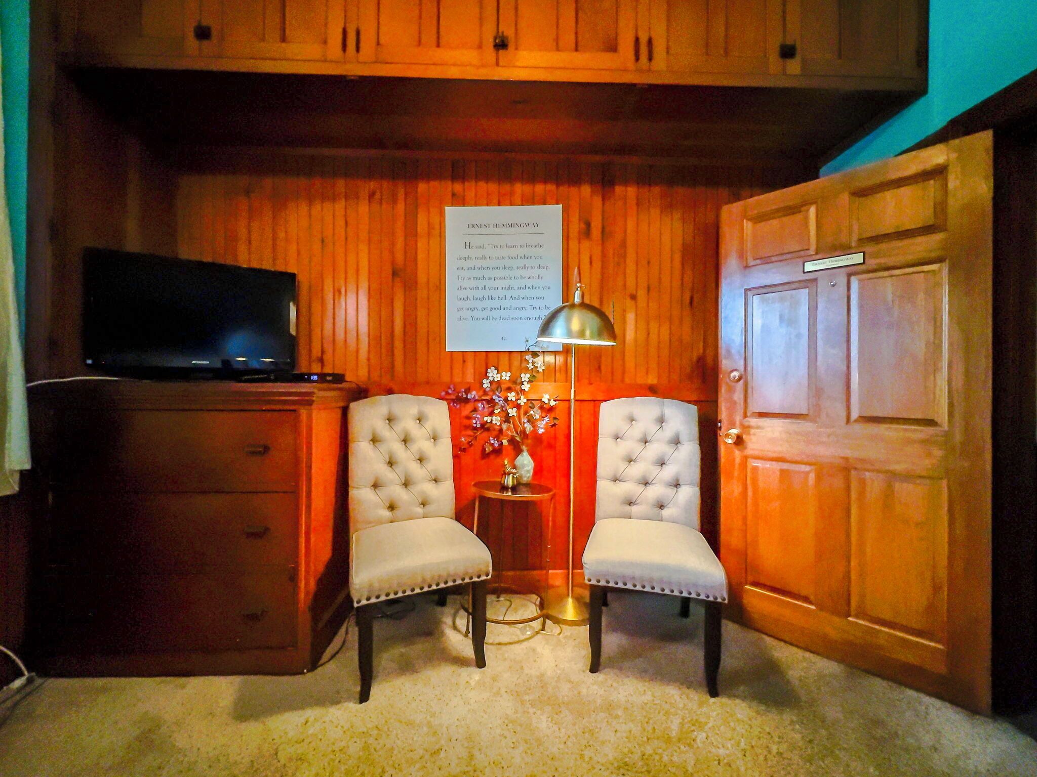 A room with wooden paneling, a television on a cabinet, two upholstered chairs, a small table with a lamp, and a door with a sign.