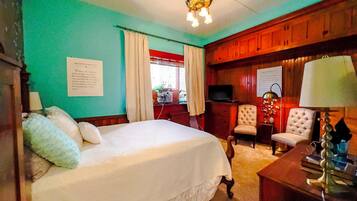 Une chambre d’hôtel comprenant un lit, deux fauteuils, un bureau avec une lampe, une télévision et un lustre.