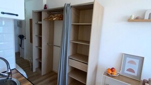 A compact kitchen with a sink and wooden cabinets, a built-in wardrobe with hanging space, and a small table with books and a basket of fruit.