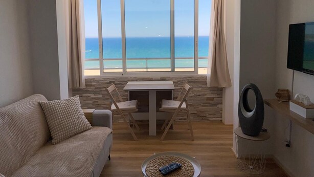 Un salón con vista al mar, un sofá, una mesa con sillas y un televisor en la pared.