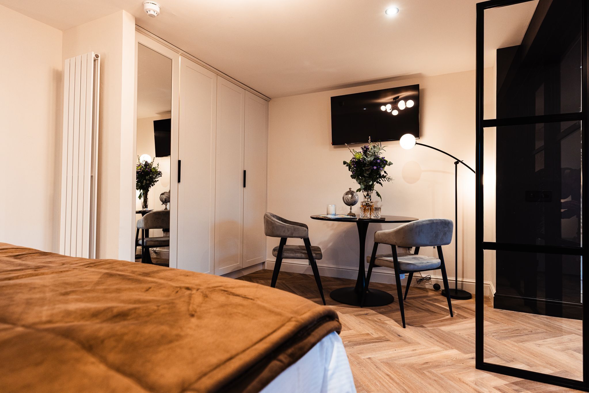 A bedroom with a bed, a desk with two chairs, a TV mounted on the wall, and a potted plant.