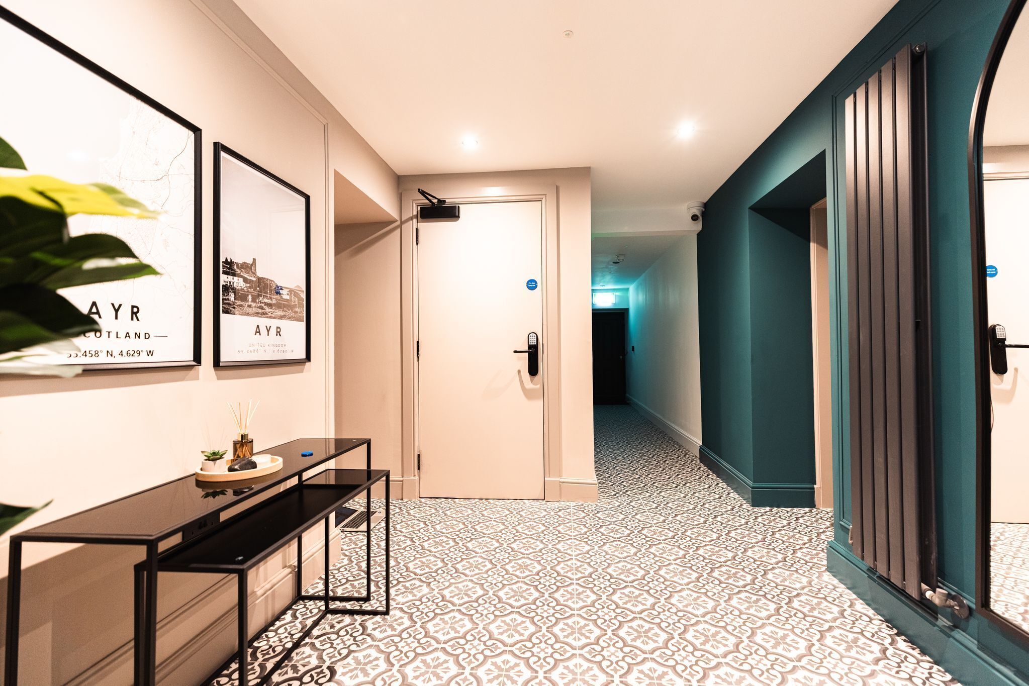 A hallway with patterned flooring, a black console table, framed pictures on the wall, and a teal-colored door.