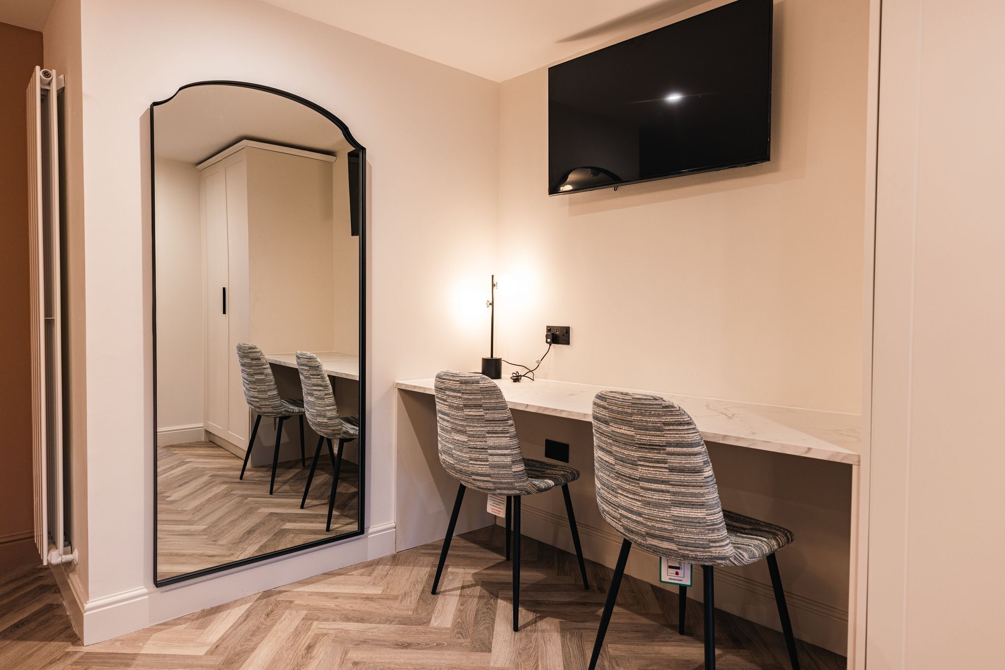 A modern room with a large mirror, a desk, two patterned chairs, a flat-screen TV mounted on the wall, and a light fixture above the desk.