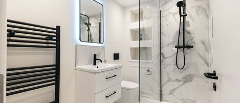 Un baño moderno con ducha envuelta en vidrio, tocador blanco con espejo y toallero negro.