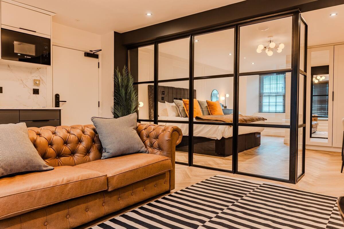 Ein modernes Wohnzimmer mit einem Ledersofa, einem Streifenmuster-Teppich und einer Schiebetür zu einem Schlafzimmer mit einem Bett und einem Nachttisch.