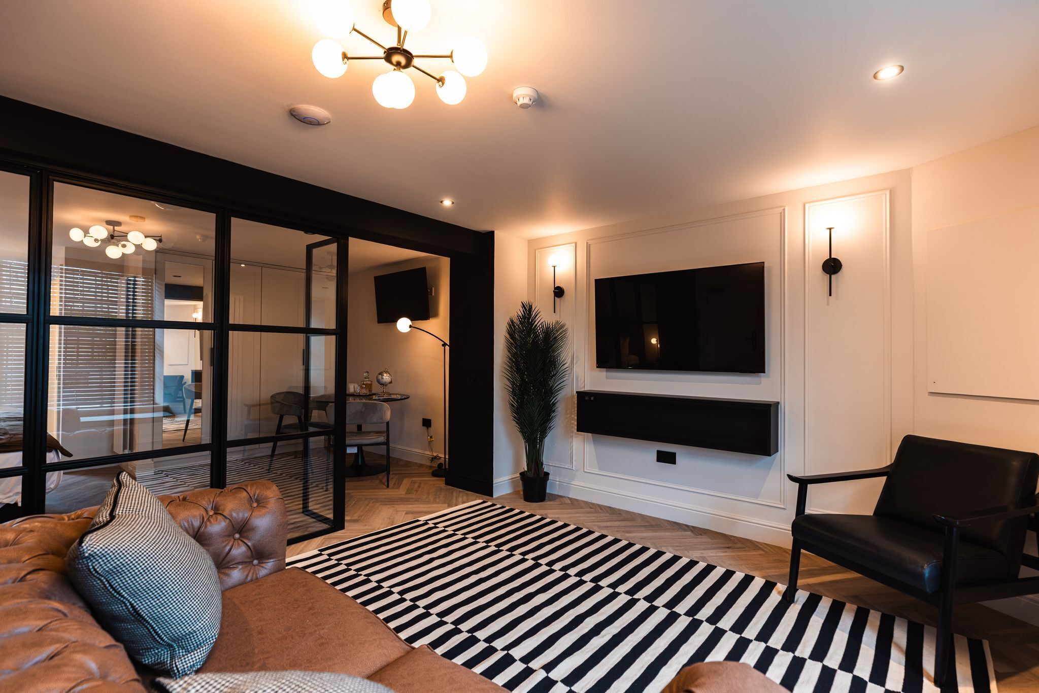 Un salón moderno con un amplio sofá modular, un tapete a rayas blanco y negro, un sillón de cuero negro, una planta en maceta y un televisor montado.