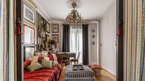 Une pièce avec un canapé rouge, un pouf à rayures, un lustre et un coin repas comprenant une table et des chaises.