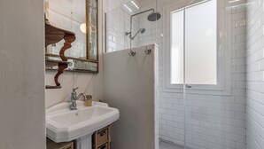 Une salle de bain avec un lavabo blanc, un miroir doté d’un cadre orné, une douche avec une porte en verre et des murs recouverts de carreaux blancs.