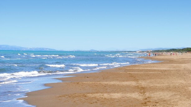 Ein weiter Sandstrand mit sanften Wellen, blauem Himmel und Bergen in der Ferne.