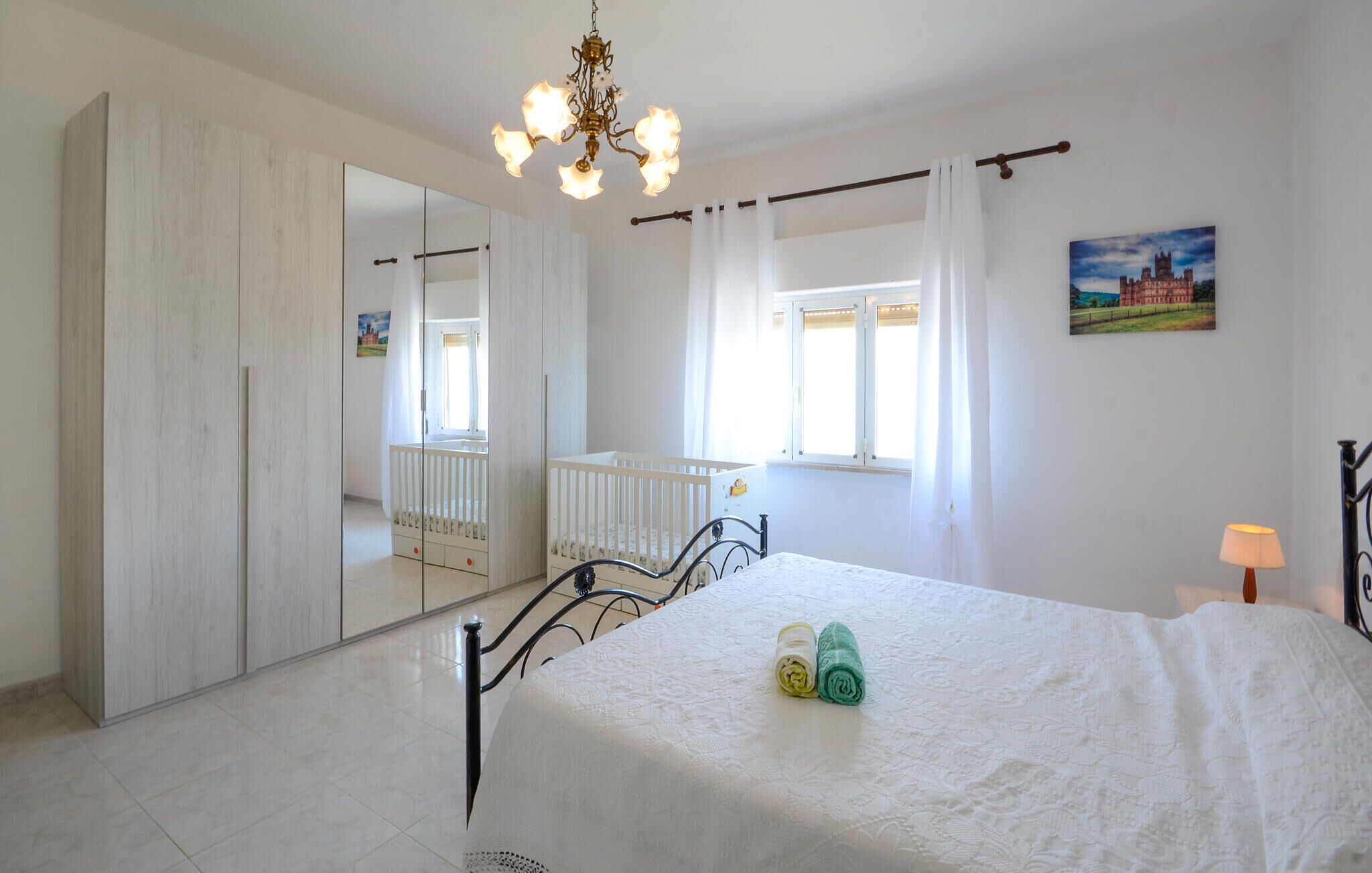 Ein Schlafzimmer mit einem großen Spiegel, einem Kinderbett, einem Bett mit gerollten Handtüchern und einer Chandelier.