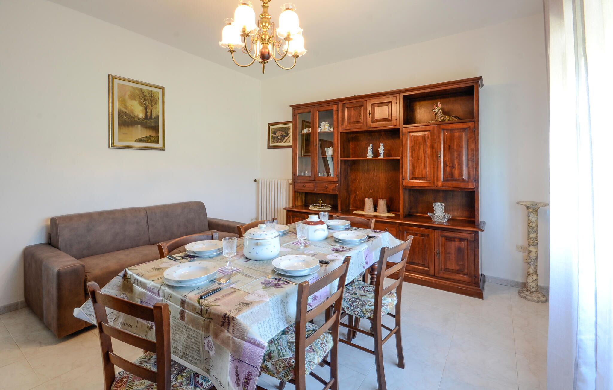 A dining area with a table set for four, surrounded by wooden chairs. A brown sofa is against the wall, and a wooden cabinet with glass doors is to the right.