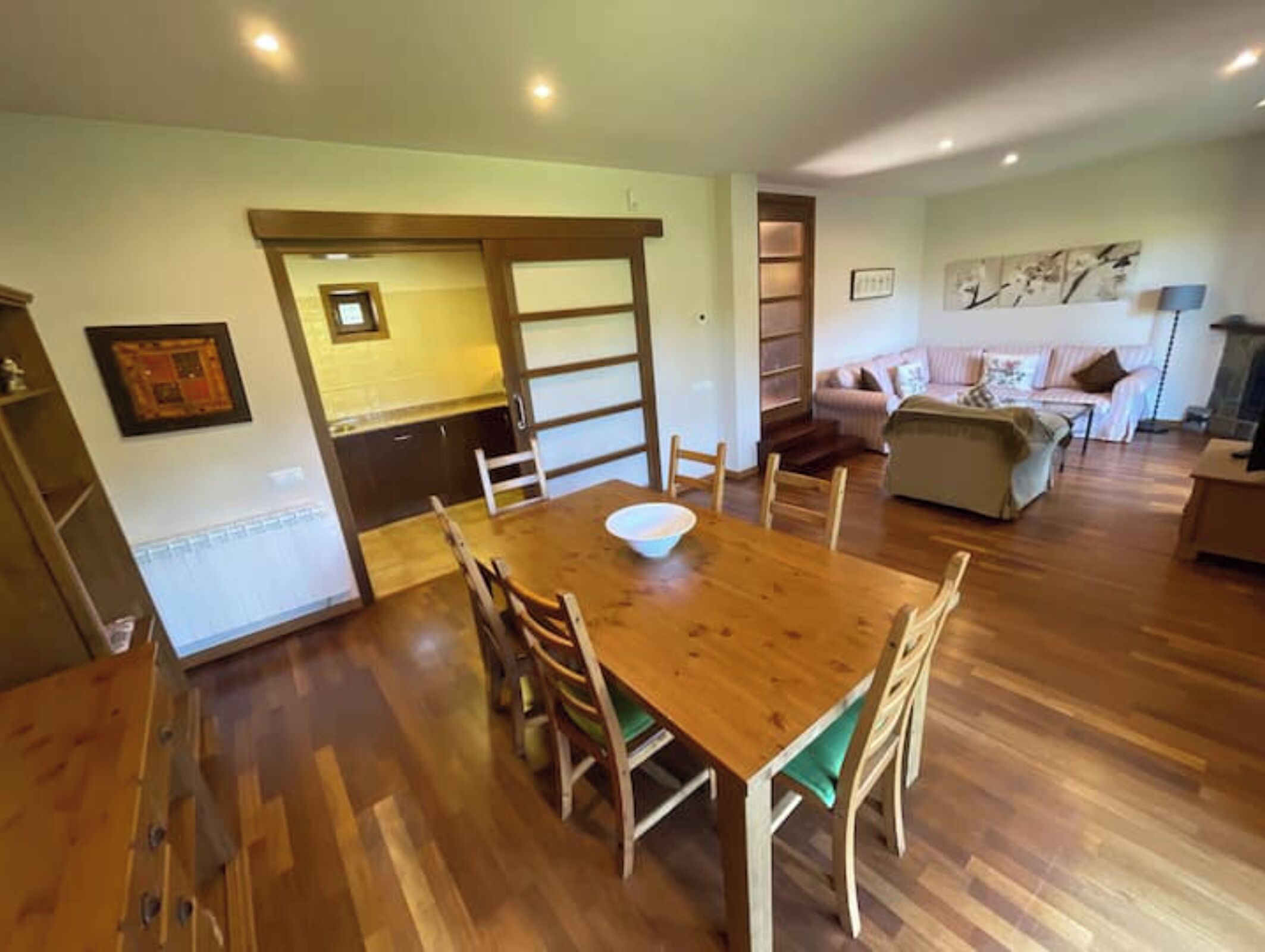 Un comedor con una mesa y sillas de madera, una cocina al fondo y una sala de estar con un sofá y chimenea.