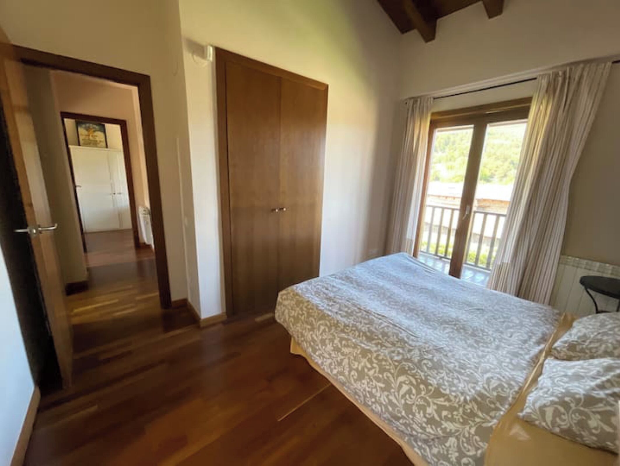 Une chambre avec un grand lit, un plancher en bois, une porte coulissante et un balcon agrémenté de rideaux.