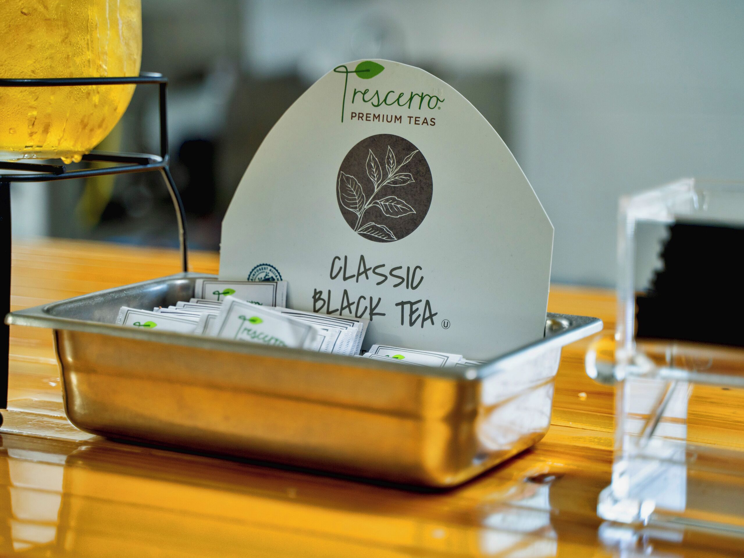 Une assiette contenant des sachets de thé étiquetés « Thés Trescero Premium, thé noir classique », avec un infuseur à thé et une tasse de thé en arrière-plan.