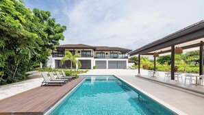 A modern pool area with a wooden deck, lounge chairs, and a covered seating area. - Luxury Beachside Condo in Hacienda Pinilla-Steps from Sand! Beach Club access (Tamarindo)