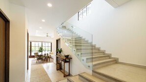 A modern interior with a glass staircase leading to a second floor. A dining area with a sofa and table is visible in the background. - Luxury Beachside Condo in Hacienda Pinilla-Steps from Sand! Beach Club access (Tamarindo)