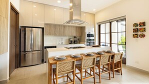 A modern kitchen with stainless steel appliances, a central island with bar stools, and large windows providing natural light. - Luxury Beachside Condo in Hacienda Pinilla-Steps from Sand! Beach Club access (Tamarindo)