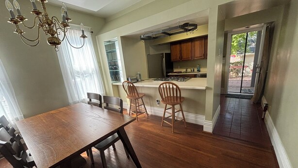 A dining area with a wooden table and chairs, a chandelier, and a kitchen with wooden cabinets and a window.