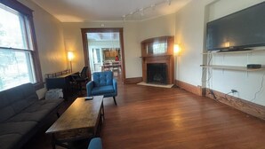 A spacious living room with a fireplace, a television mounted on the wall, and a dining area visible through an open doorway.