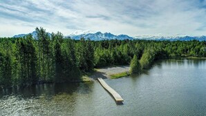A serene lakeside view with a wooden dock extending into the water, surrounded by dense forest and snow-capped mountains in the background.