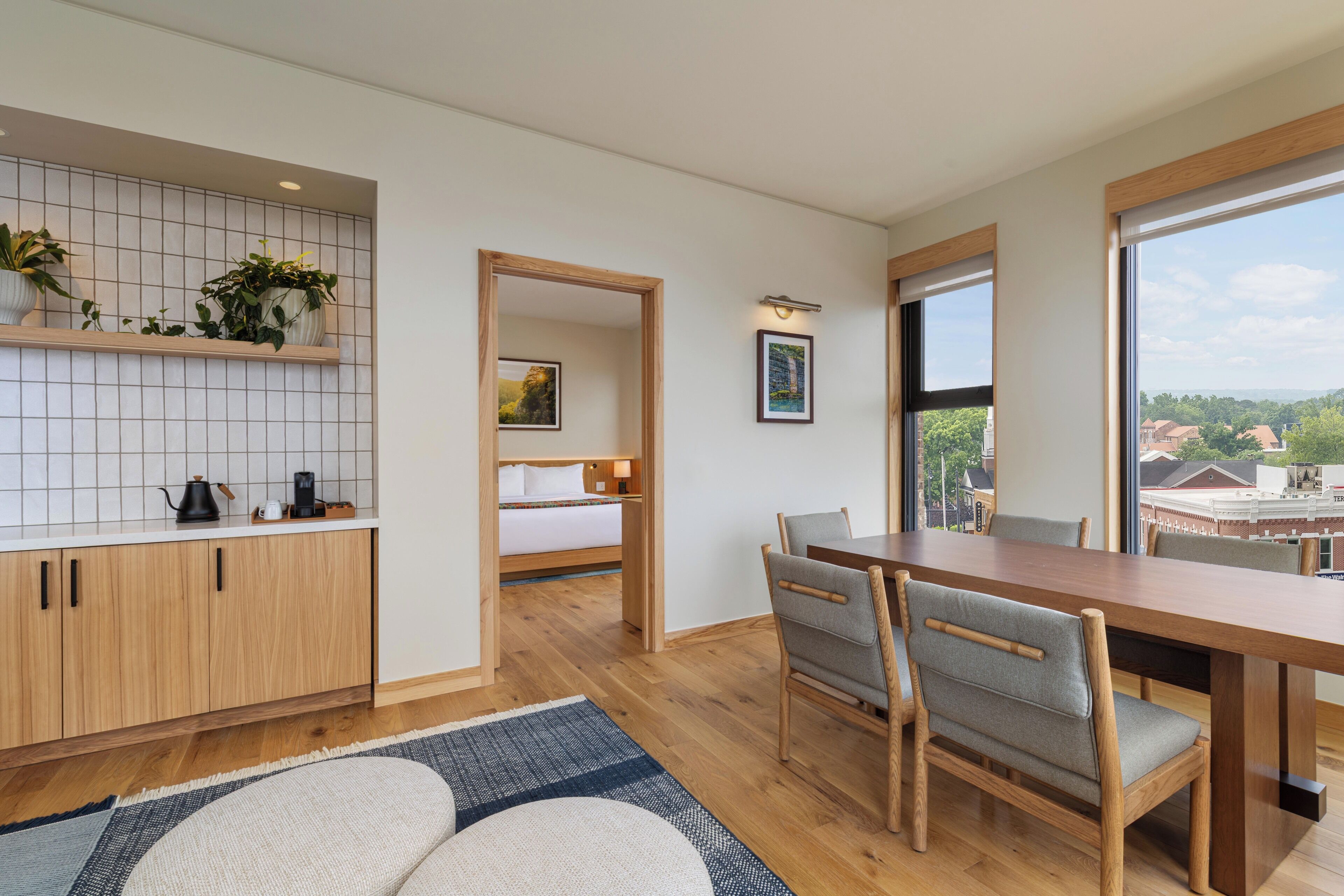 A modern hotel room with a kitchen, dining area, and a large window offering a city view.