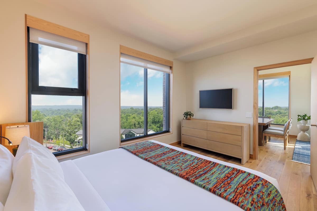 A modern hotel room with a large bed, a TV, a dresser, and a view of the forest.