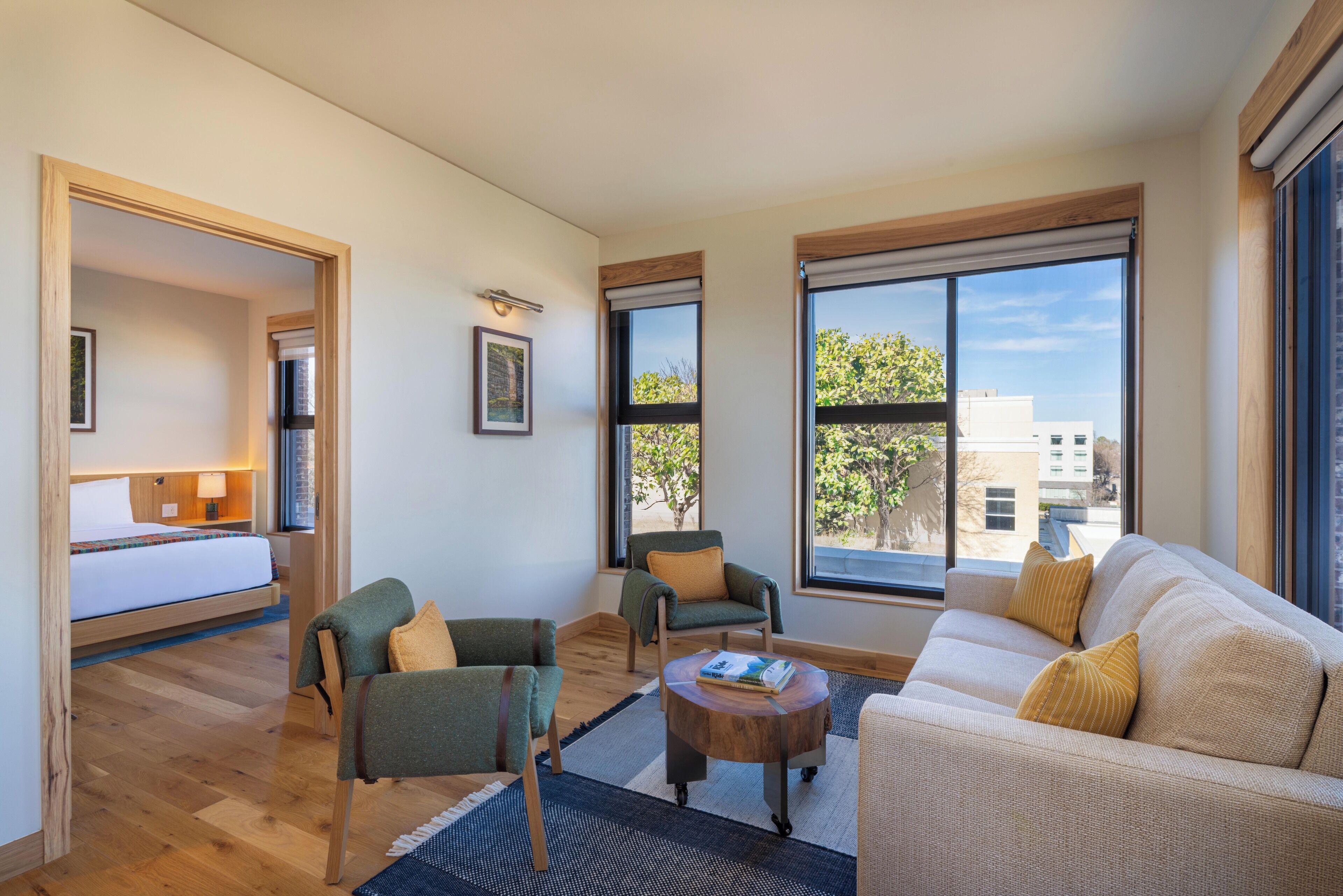 A modern hotel room with a bed, two armchairs, a sofa, a small coffee table, and large windows offering a view of the outside.