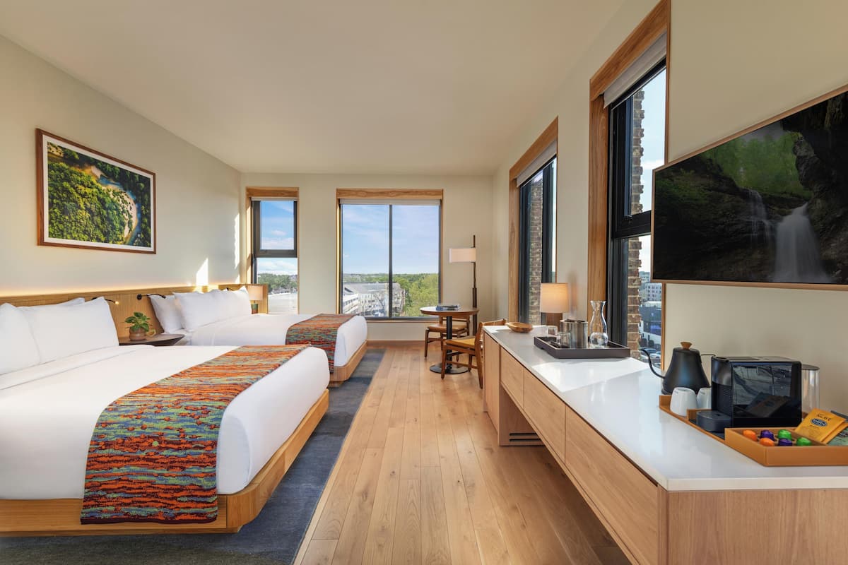 A hotel room with two beds, a desk, a TV, and large windows.