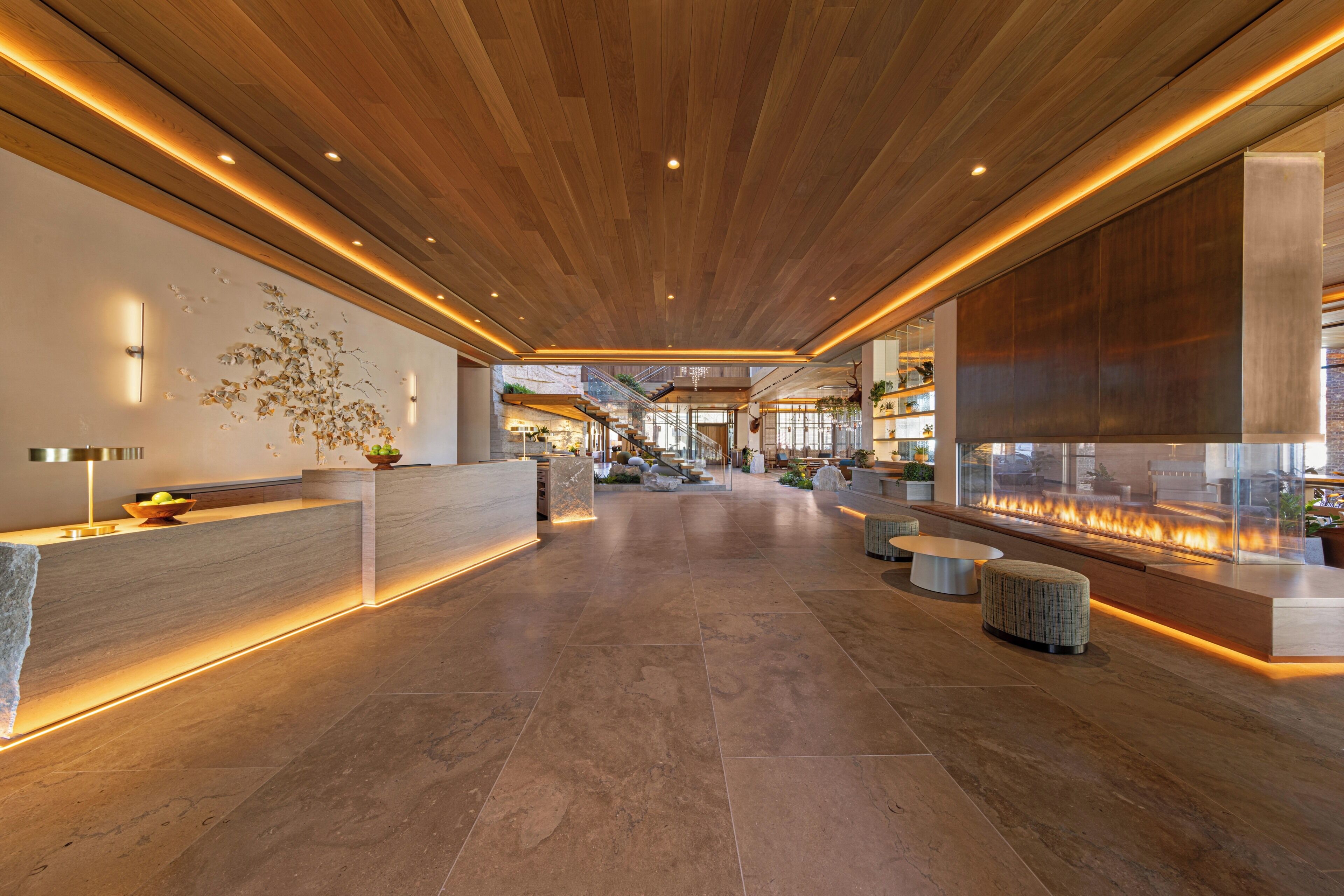 A modern lobby with a wooden ceiling, a reception desk, a fire feature, and a seating area.
