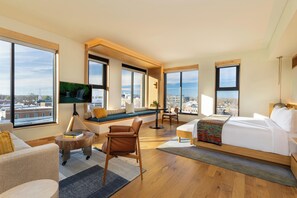 A modern hotel room with a large bed, seating area, TV, and large windows offering city views. - The Compton (Bentonville)
