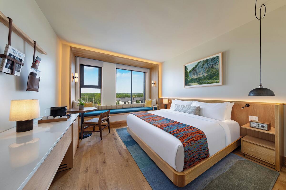 A modern hotel room with a large bed, a desk, a chair, a window with a view, and a bedside table with a lamp.