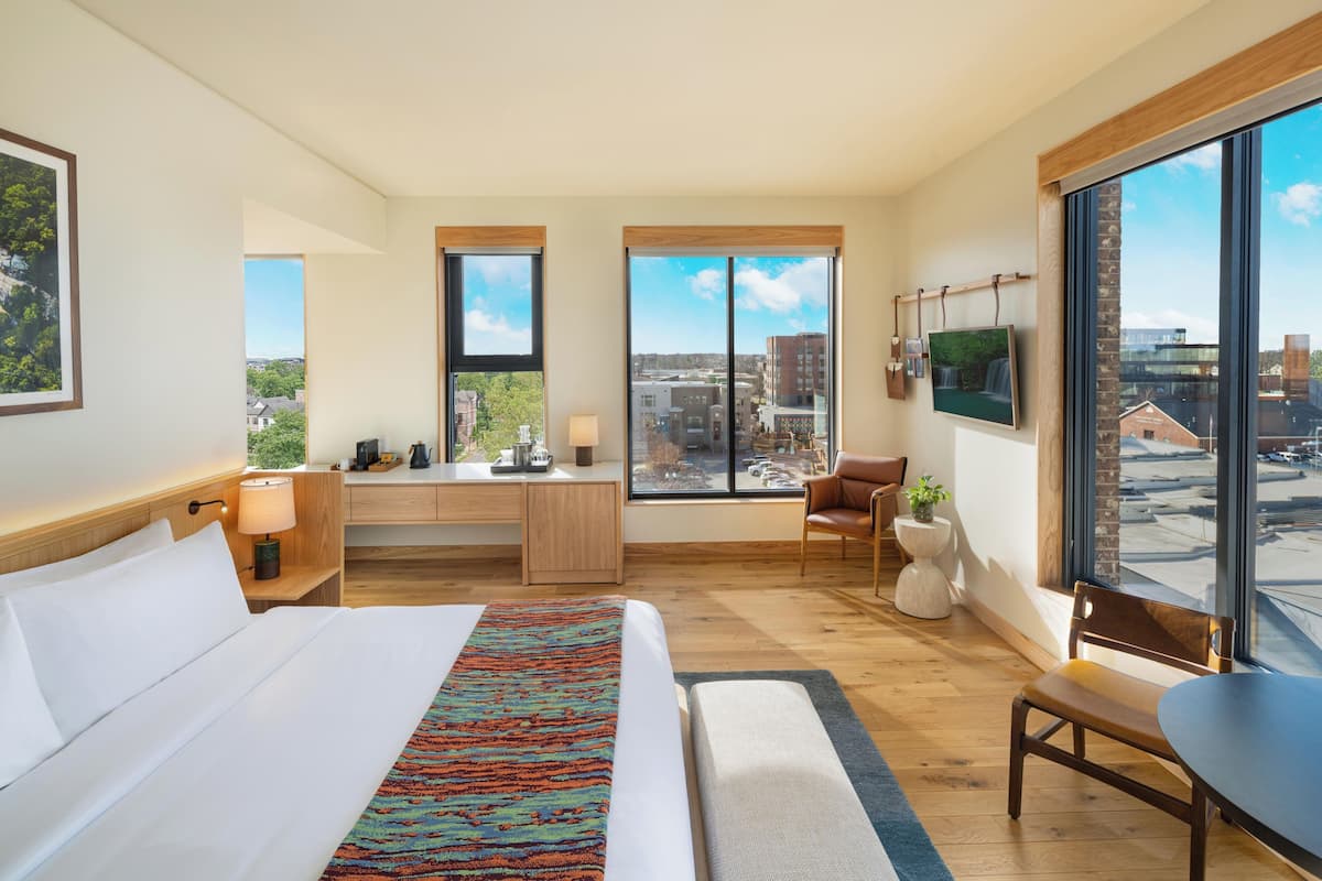 A hotel room with a large bed, a desk, a chair, a TV, and large windows offering a city view.