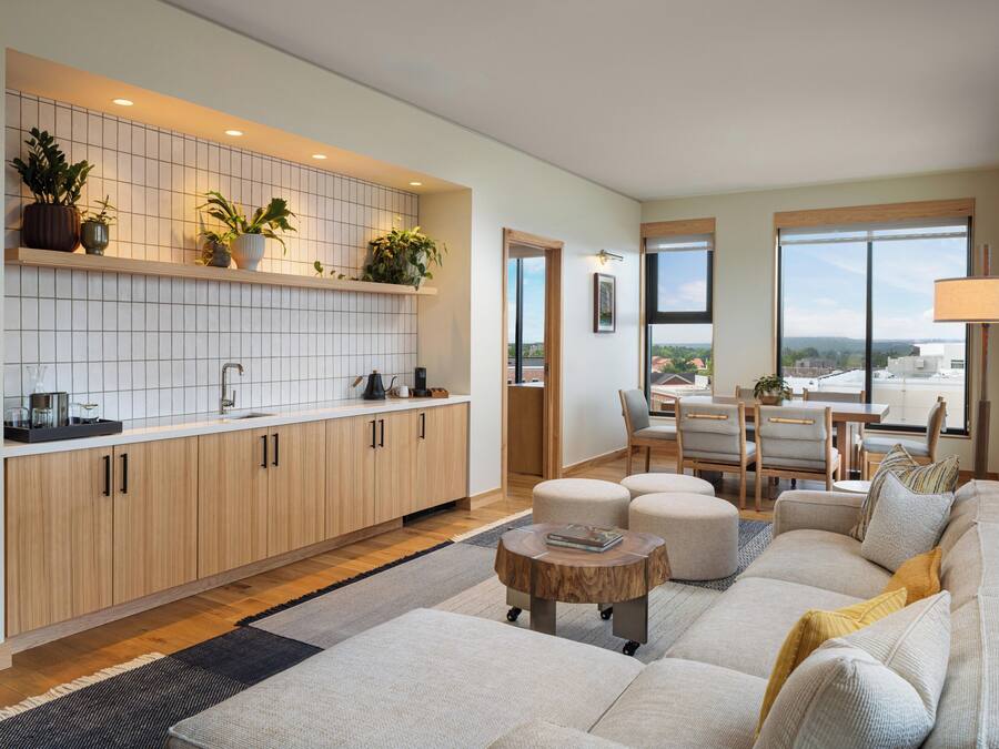 Una cocina moderna con azulejos blancos, armarios de madera y plantas en macetas. Hay una zona de comedor con una mesa y sillas, y un amplio ventanal con vistas.