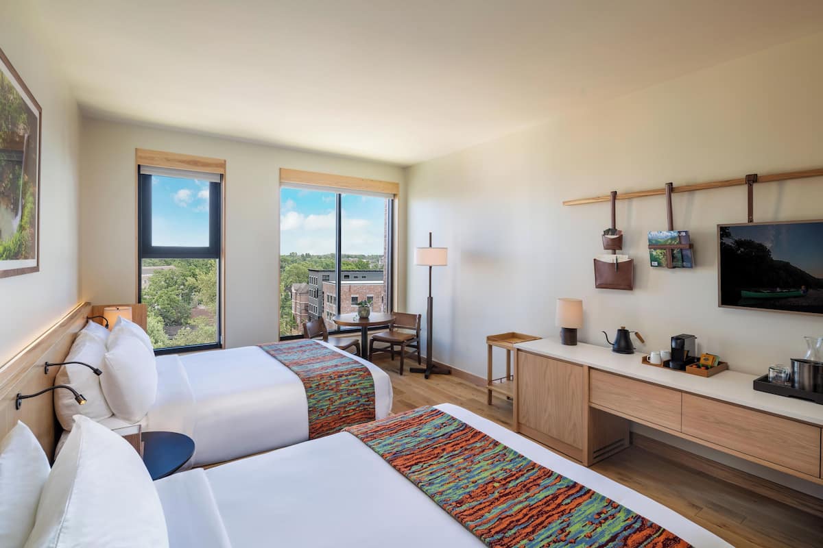 A hotel room with two beds, a desk, a chair, a TV, and a window with a view of buildings and trees.